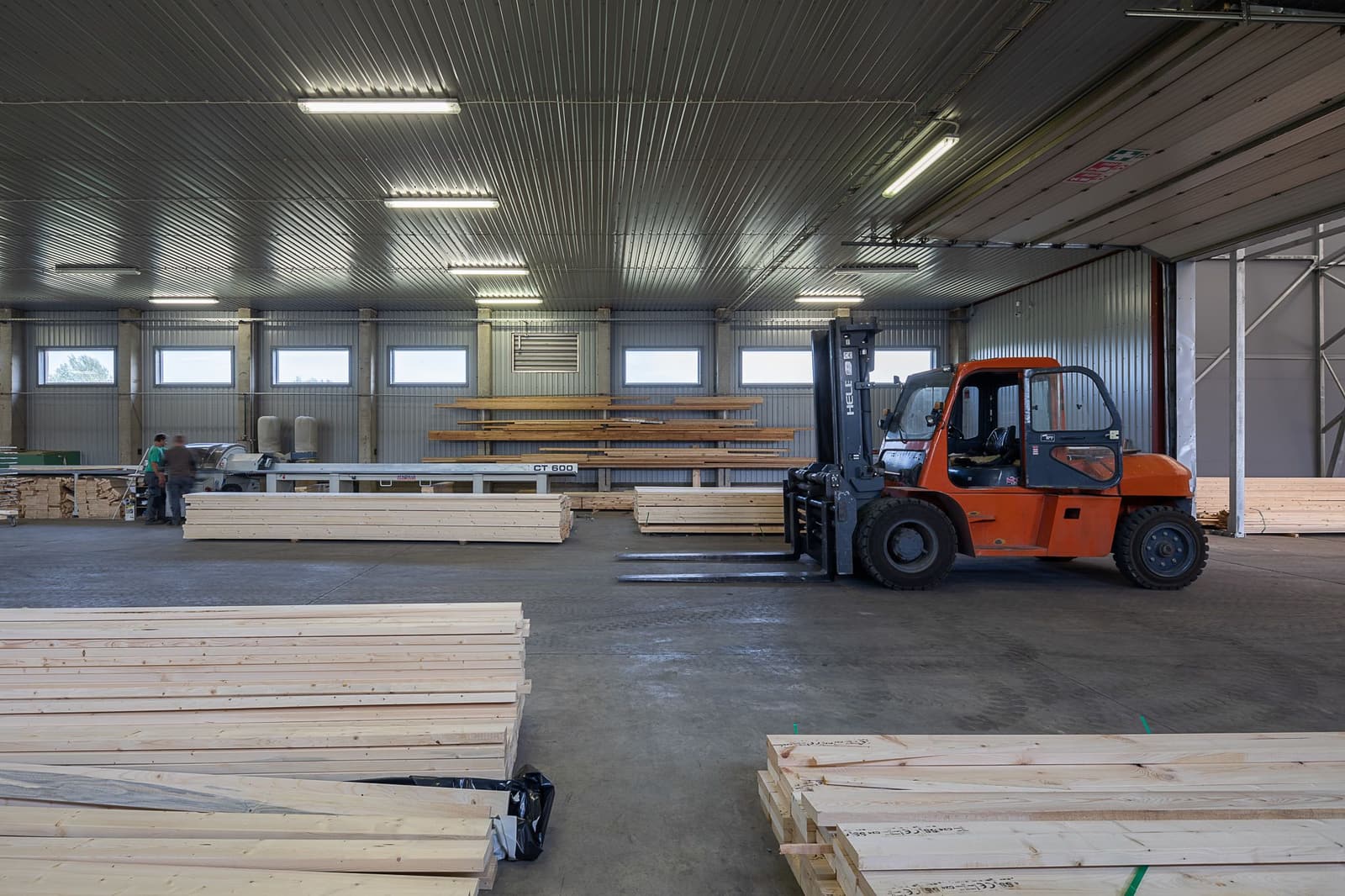 A heavy forklift moves long glulam beams through the factory hall past stacks of structural timber