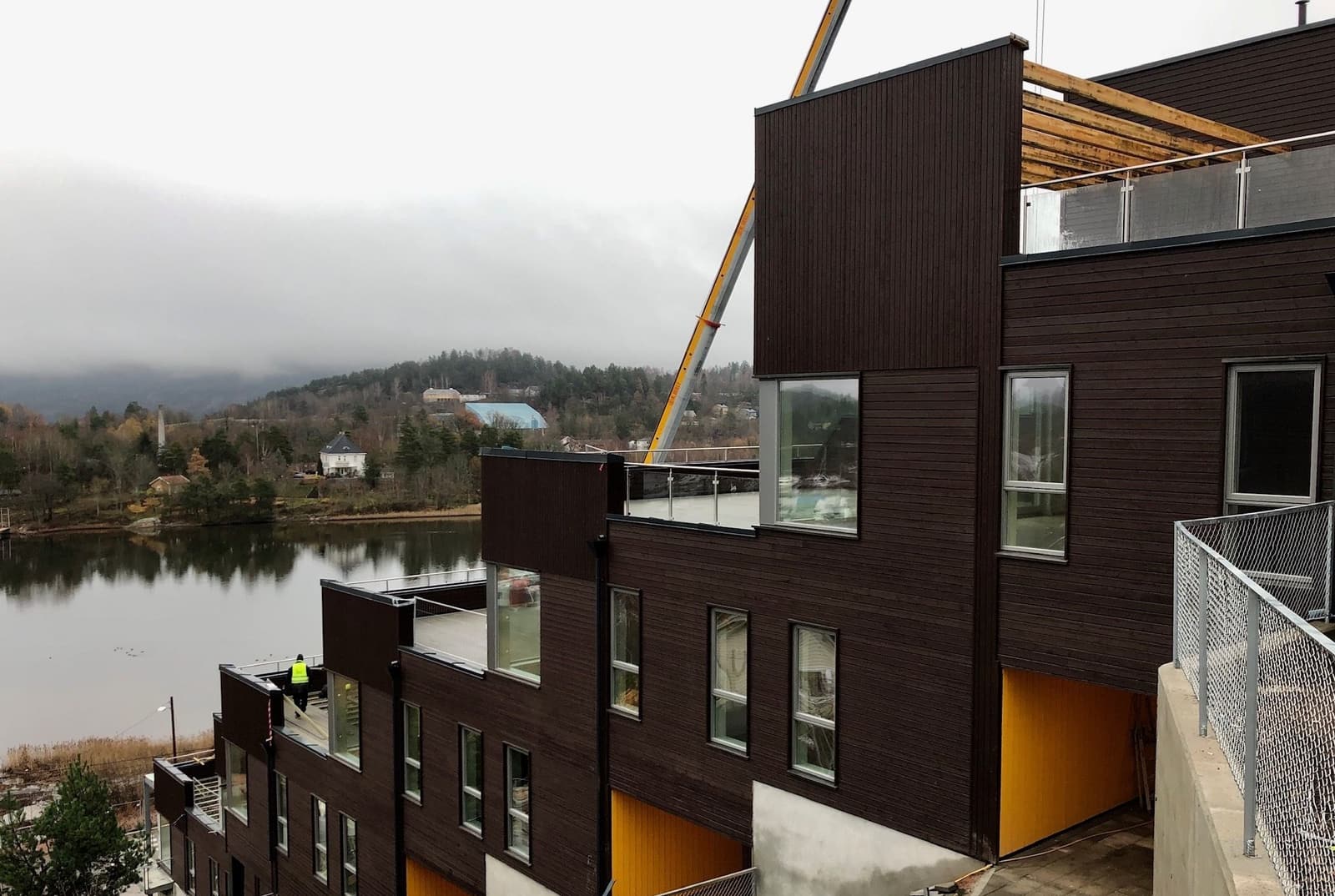A mobile crane lifts roof elements onto a terrace of dark timber-clad houses beside a lake during installation
